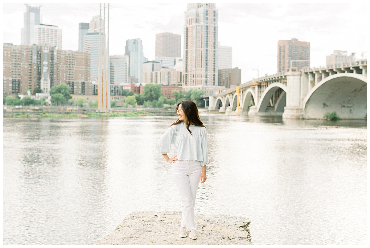 Girl in light blue top standing on a rock in front of the stone arch bridge and the Minneapolis skyline.