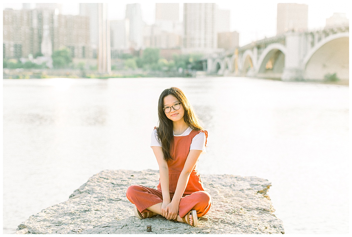 Girl in an orange jumpsuit sitting in front of the stone arch bridge and Minneapolis skyline.