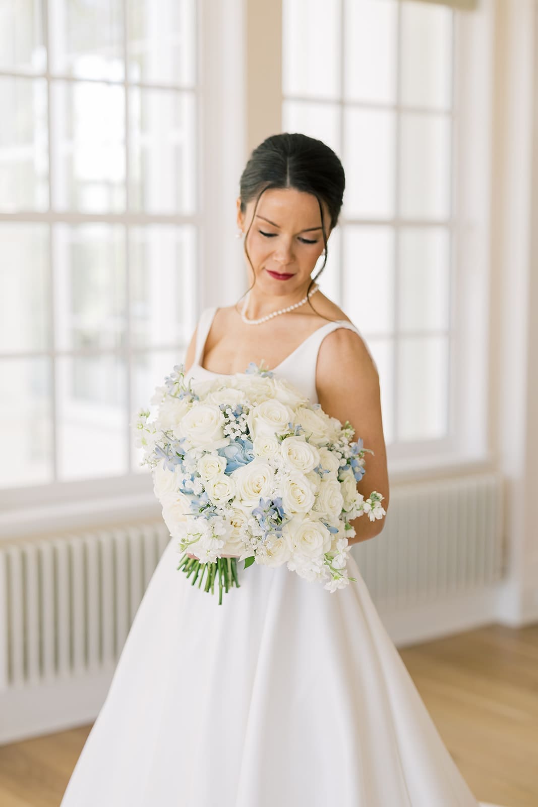 Bride looking down at her white and blue wedding bouquet of roses and hydrangeas with pearl necklace and red lip