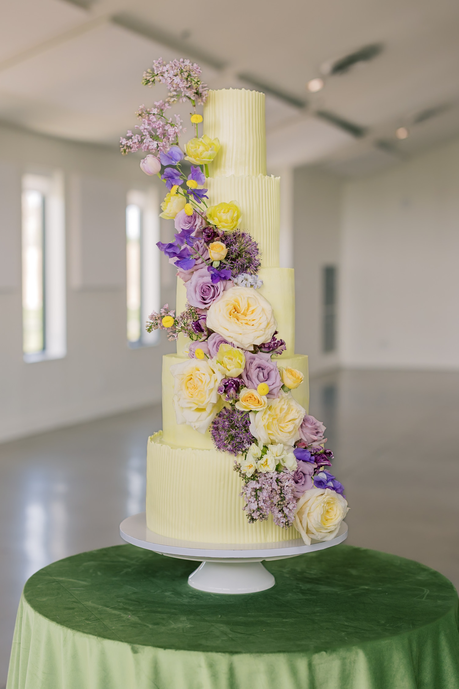 Five-tier yellow ribbed wedding cake with cascading lavender roses, purple sweet peas, and yellow ranunculus on a green velvet cake table