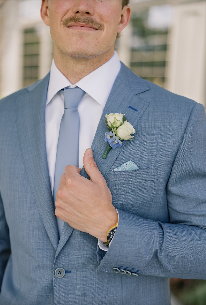 Groom smiling in a steel blue suit with a white rose and blue delphinium boutonniere, photographed outside Rush Creek Golf Club in Maple Grove MN