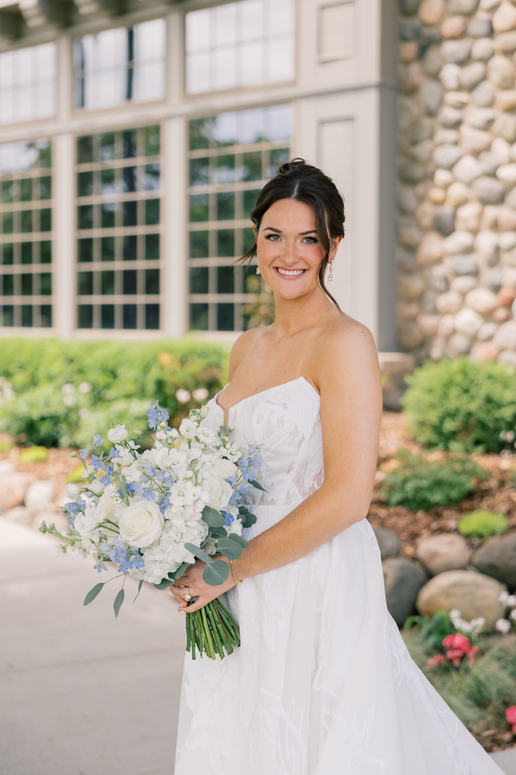 Bride smiling and holding a white and blue bridal bouquet in front of the stone and glass exterior at her Minnesota wedding venue