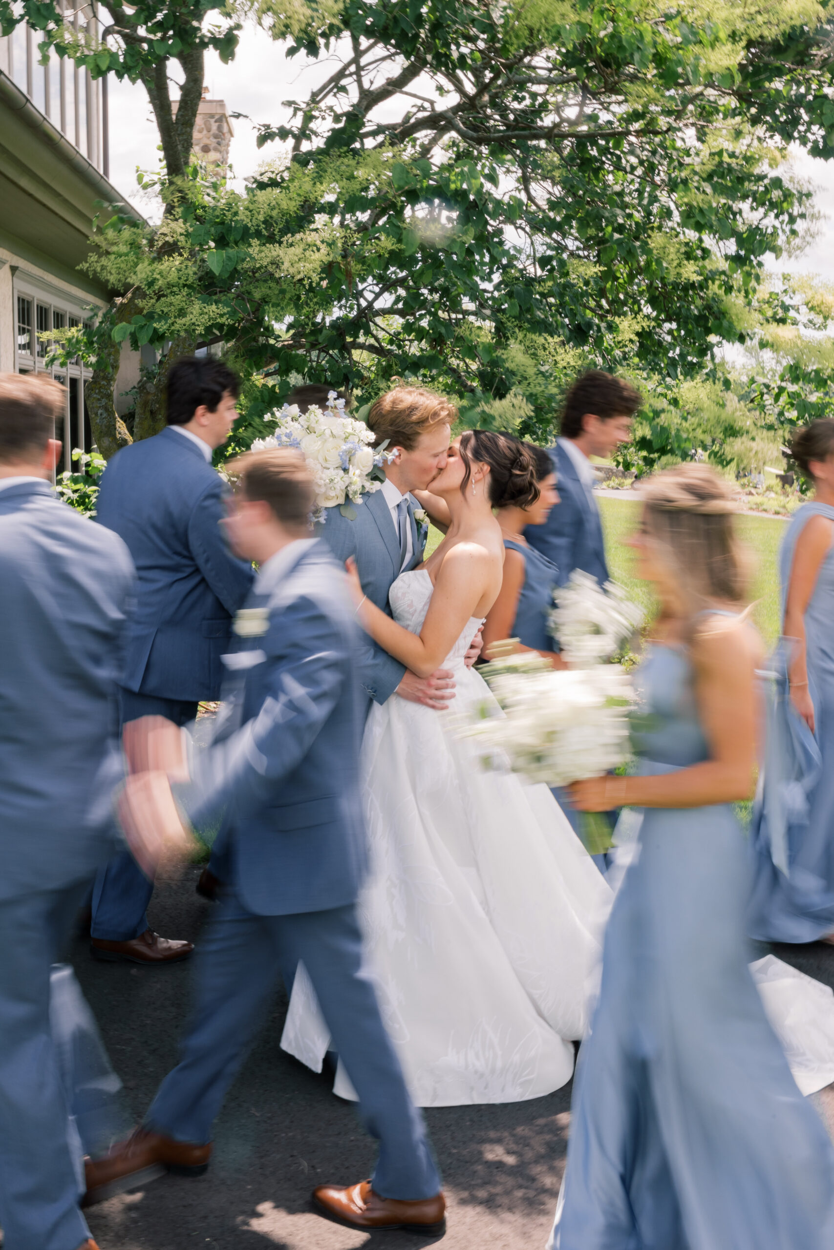 Bride and groom kissing as their wedding party in steel blue dresses and suits moves around them during portraits at their Rush Creek Golf Club summer wedding