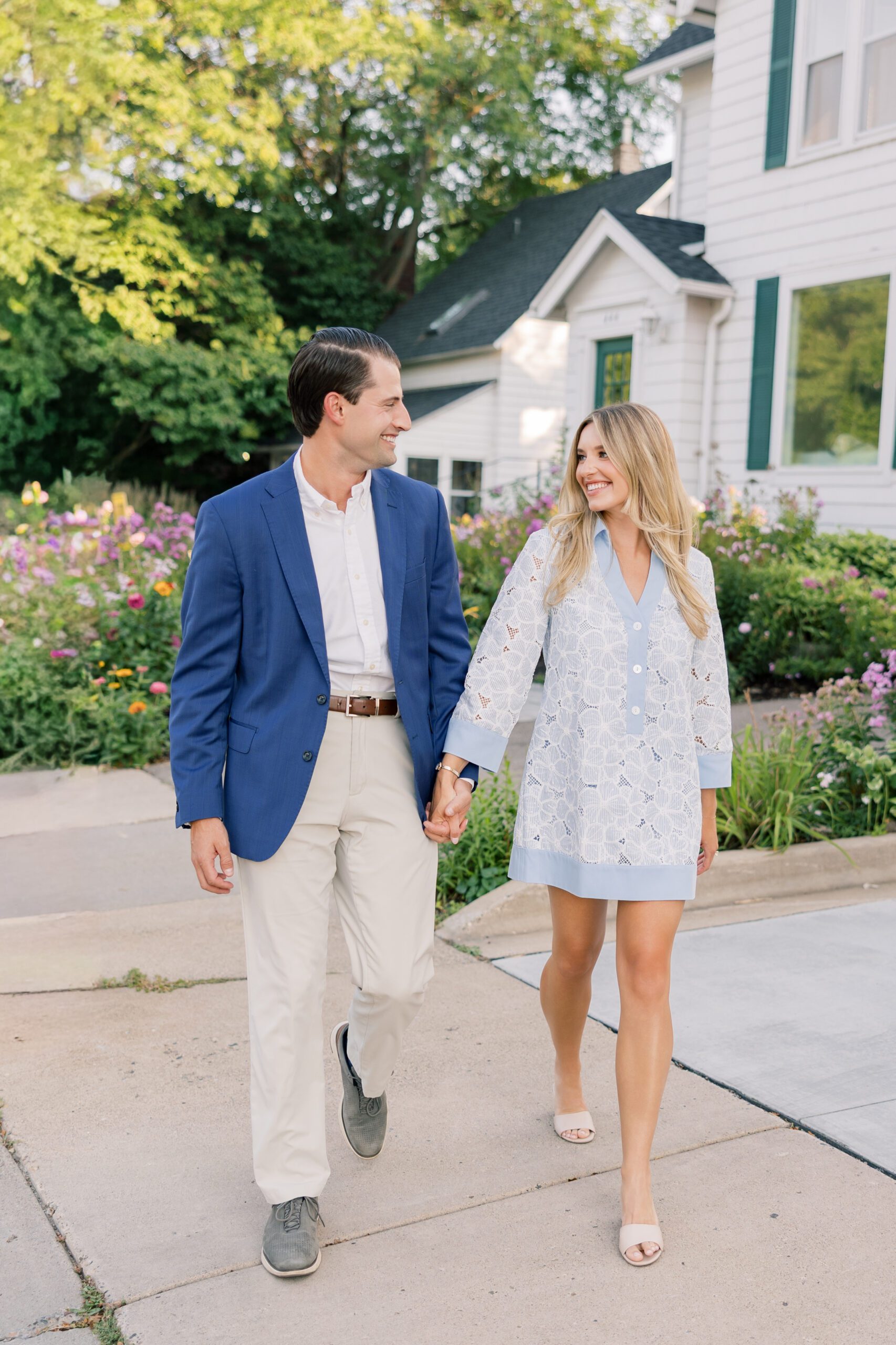 Couple walking hand in hand on a sidewalk in front of a white house with a colorful summer flower garden in Excelsior, Minnesota, showing what to wear for engagement photos: a light blue eyelet dress from Tuckernuck and a coordinating navy blazer with khaki pants