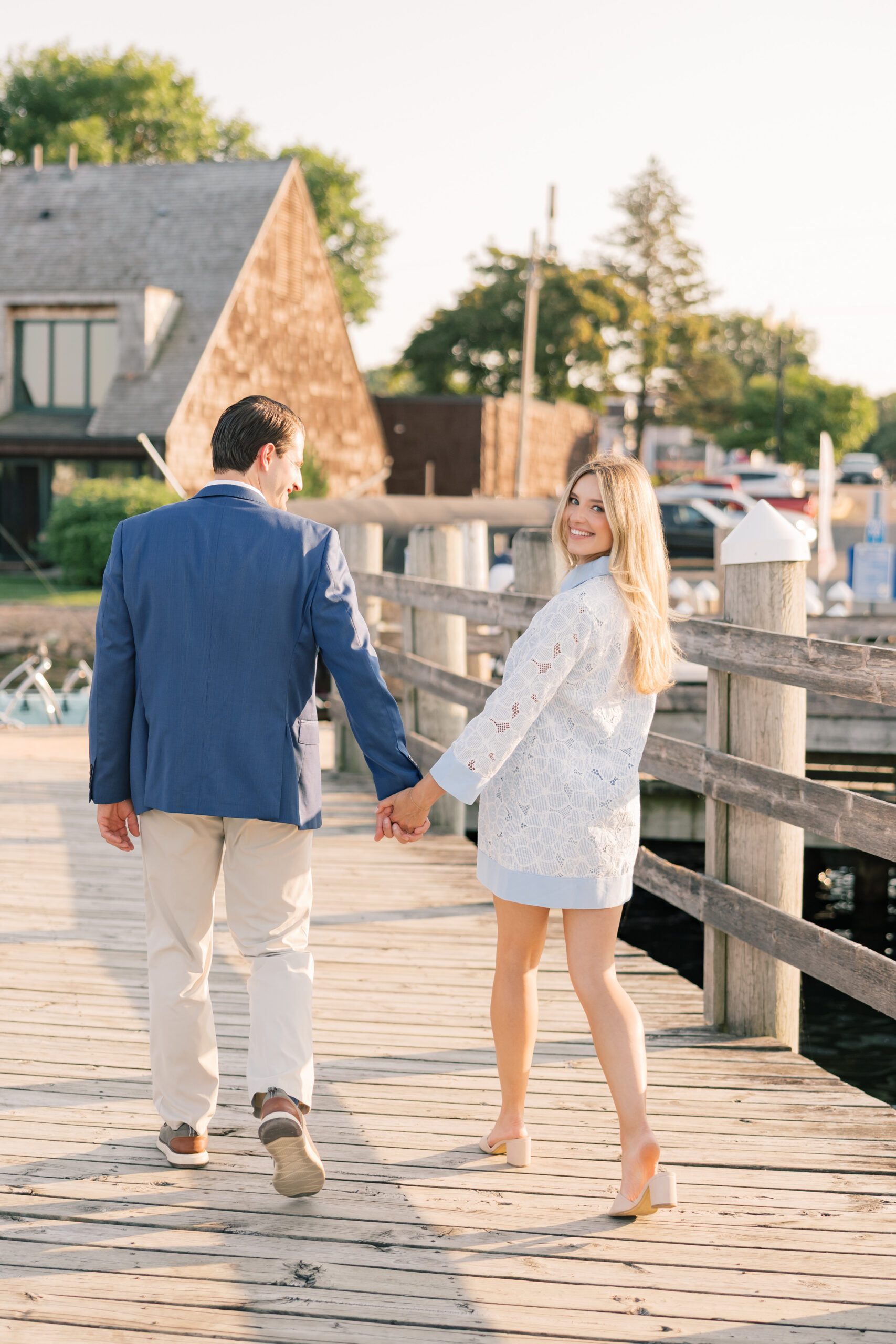 Engaged woman glancing back over her shoulder with a smile as she walks away holding her partner's hand on a dock in Excelsior, Minnesota, summer golden hour engagement session