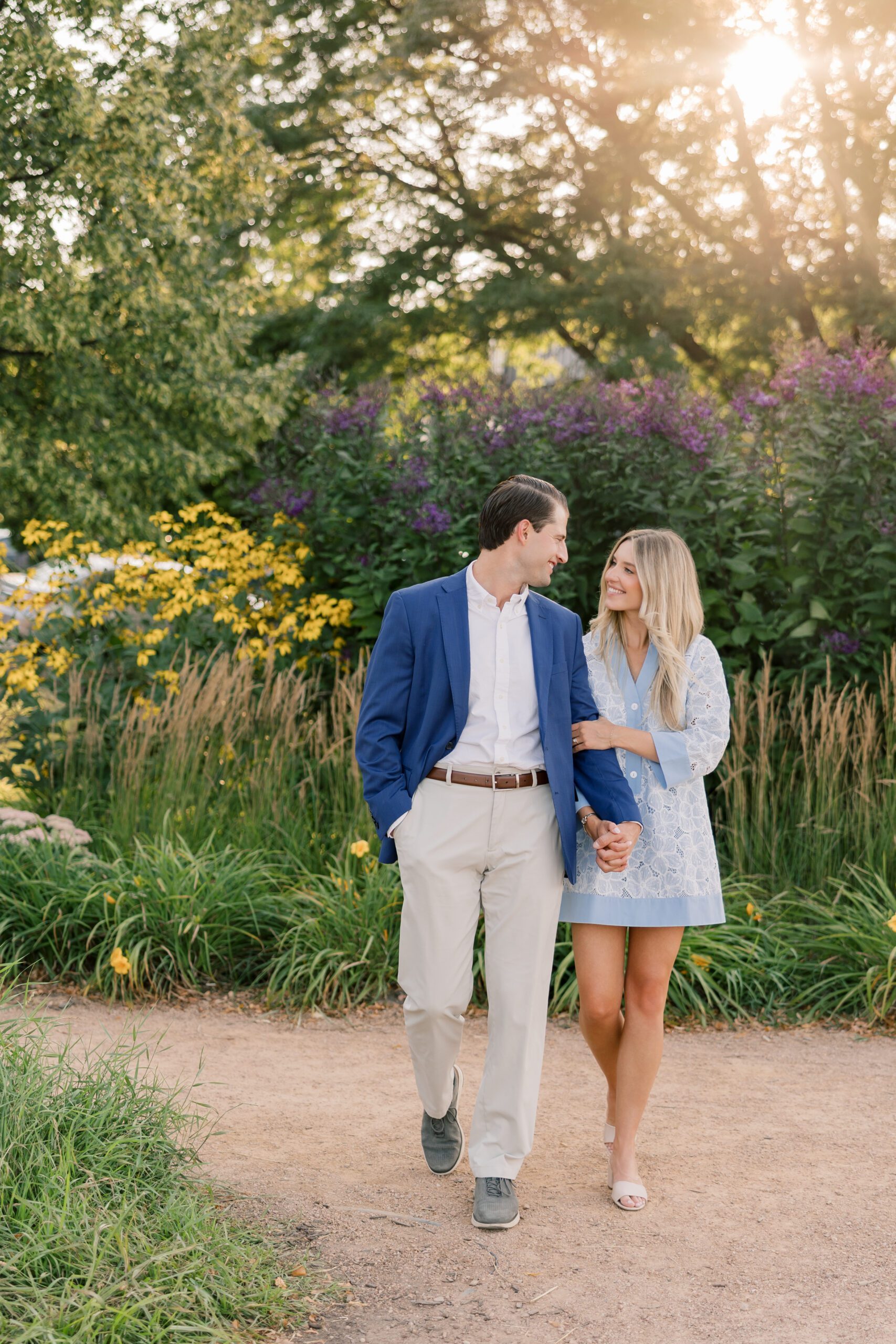 Couple walking arm in arm on a natural garden path surrounded by wildflowers and tall grasses, golden sunlight filtering through the trees, showing what to wear for engagement photos in a timeless blue and neutral color palette