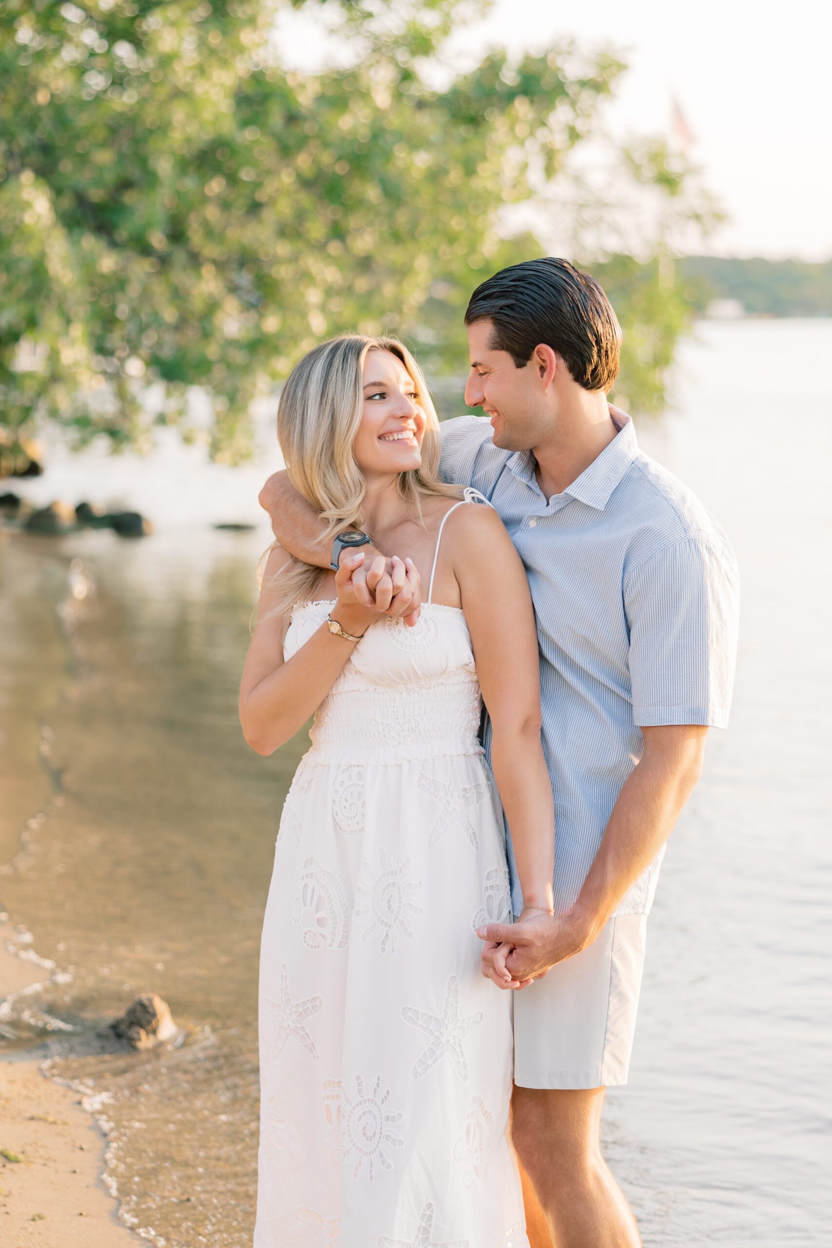 Engaged couple standing together on a sandy Minnesota lakeshore at golden hour, showing what to wear for engagement photos with a casual second look: a white lace spaghetti strap sundress and a light blue seersucker short-sleeve shirt with khaki shorts