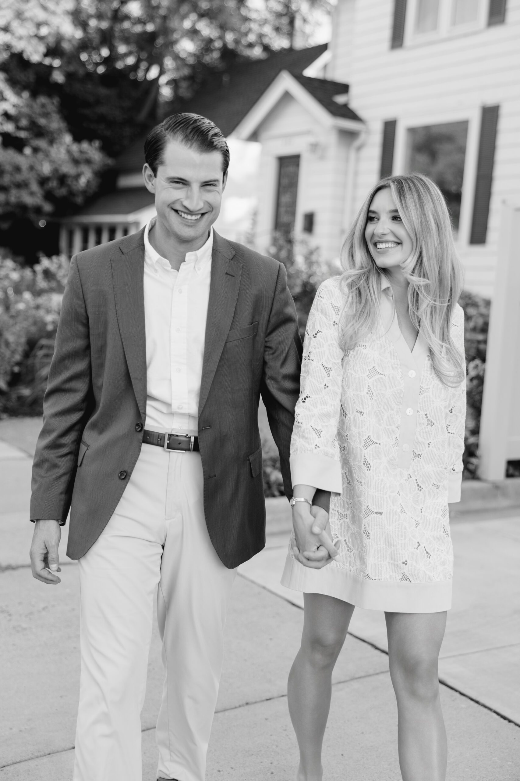 Black and white engagement photo of a couple smiling and walking together past a white house with green shutters, wearing a classic eyelet dress and blazer combination, Excelsior MN engagement session