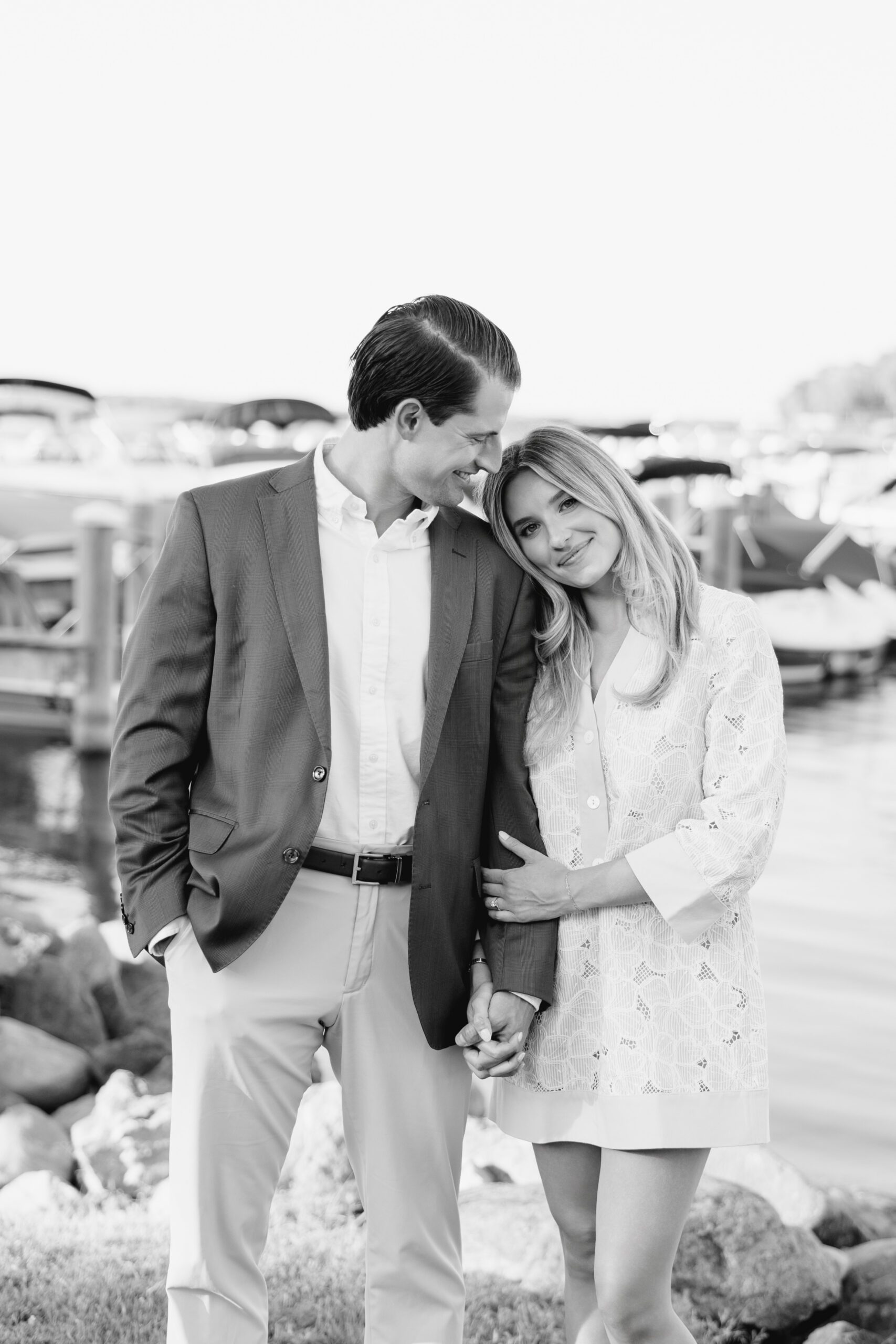 Black and white photo of an engaged couple standing close together at a lakeside marina in Excelsior, Minnesota, she leans her head on his shoulder, both wearing classic neutral and blue outfits