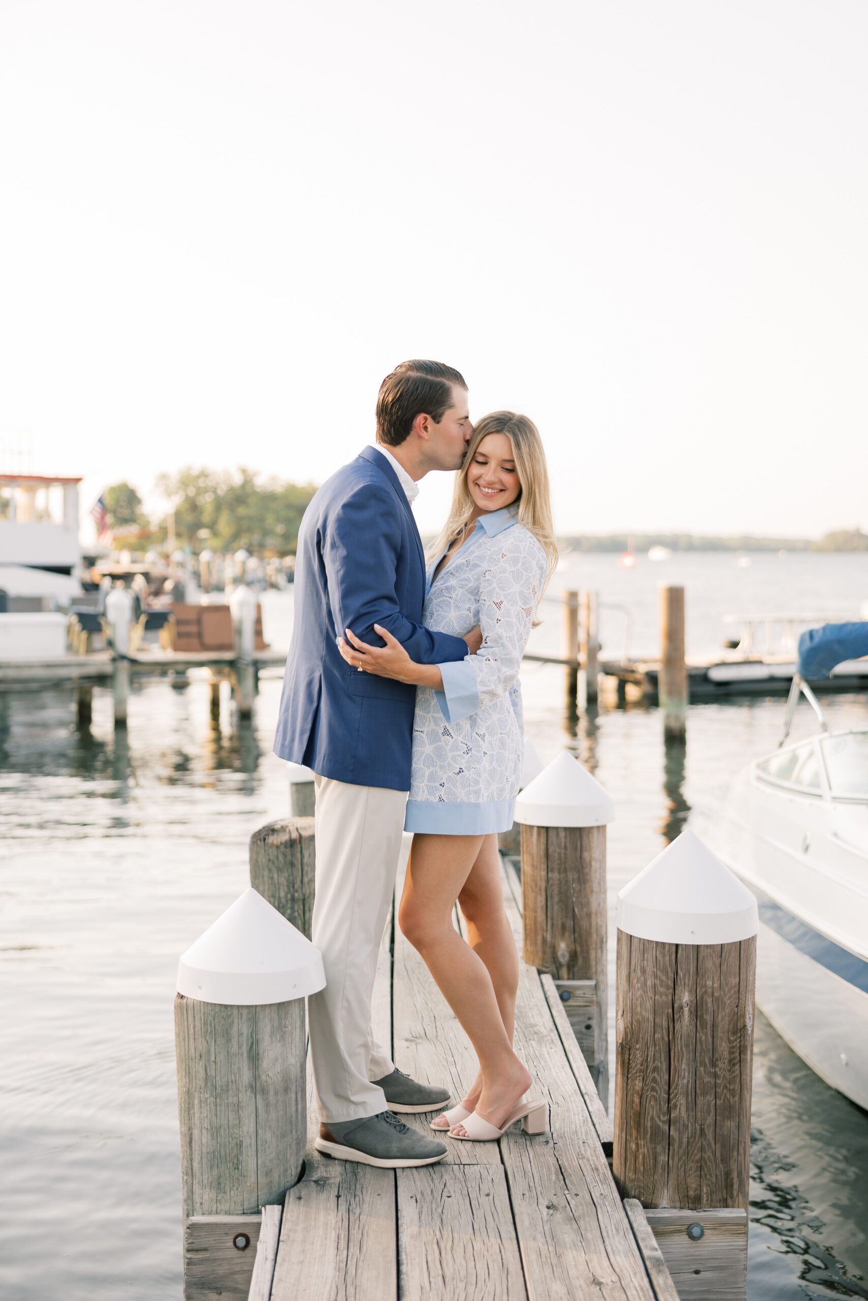 Groom kissing his fiancée on the forehead on a wooden dock at golden hour, boats visible in the background on Lake Minnetonka, light blue eyelet dress and navy blazer engagement session
