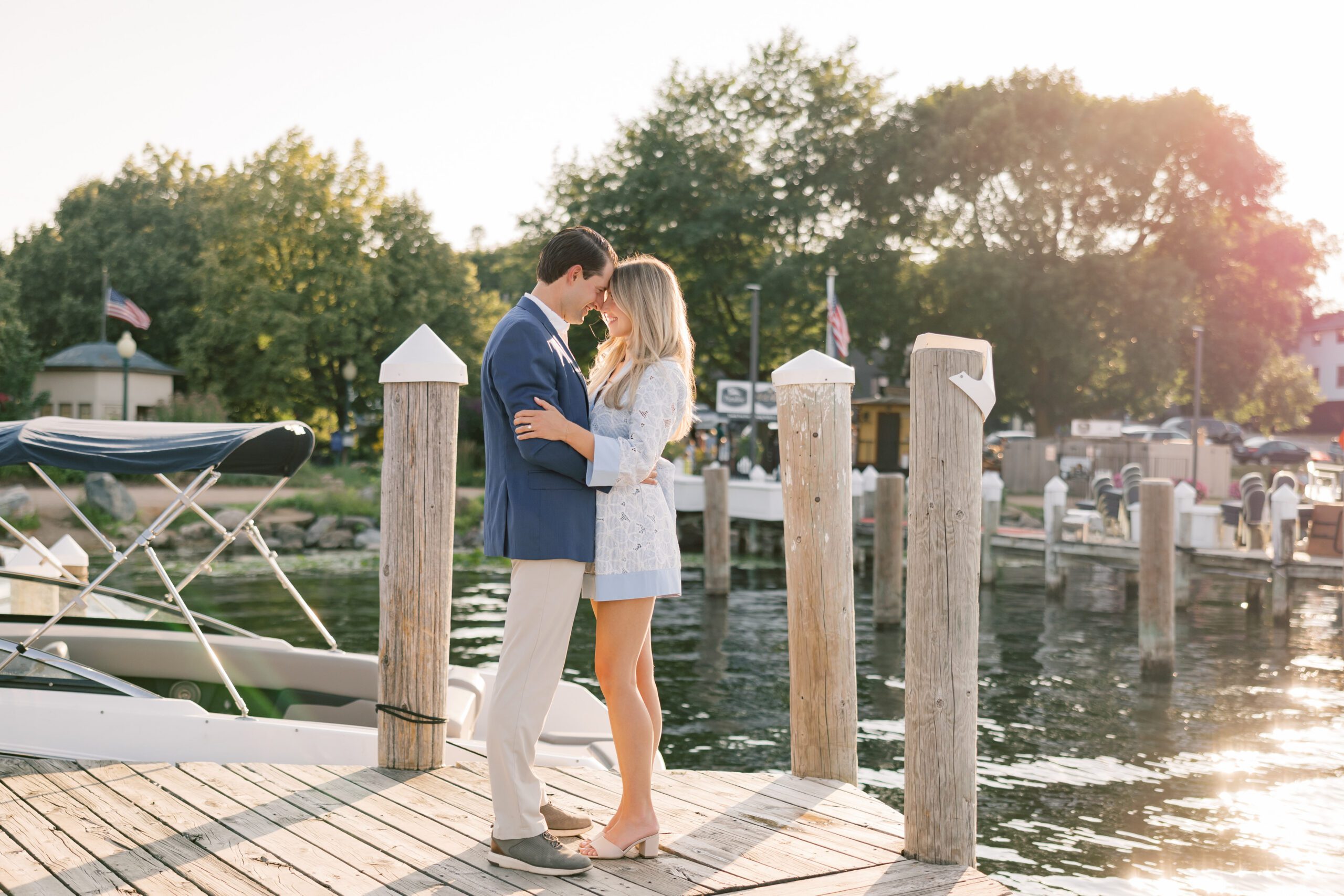 Couple facing each other with foreheads almost touching on a marina dock at sunset, soft golden light reflecting off the water behind them, summer engagement session in Minnesota